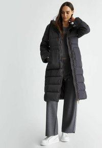 Lunga giacca invernale nera con zip frontale, cappuccio e dettagli dorati, abbinata a pantaloni da lounge grigi e sneakers bianche. Materiale liscio e isolante.