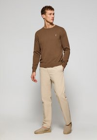 Bruine gebreide trui met een ronde hals, gecombineerd met lichtbeige broek en beige instappers, wat zorgt voor een casual outfit.