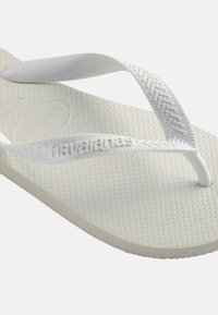 Chancla Havaianas blanca con plantilla texturizada y correas de goma con patrón que presentan el nombre de la marca en relieve en el costado.