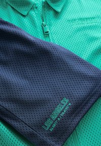 Grön perforerad tyg kortärmad polo med marinblå ärm, som har en broderad "LOS ANGELES" detalj i grönt. Dragkedja framtill.