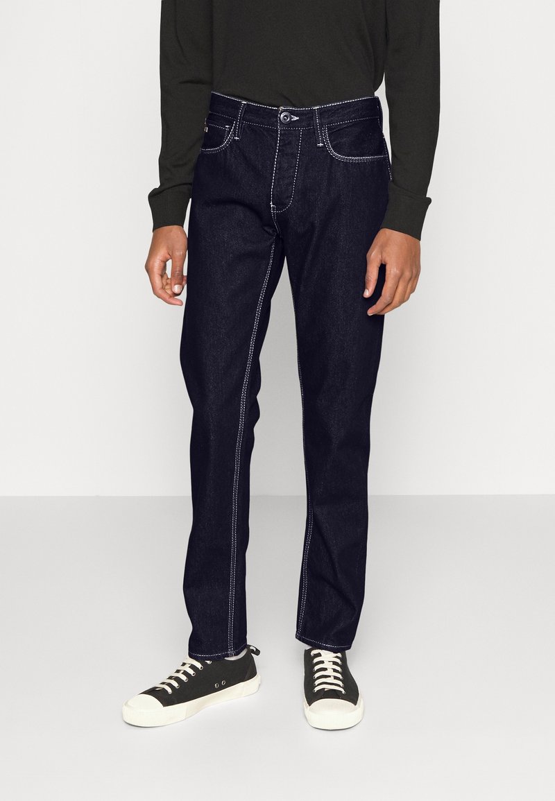 Jeans de mezclilla azul oscuro con costuras en contraste blanco, corte recto y estilo de cinco bolsillos. Combinados con zapatillas negras y blancas.