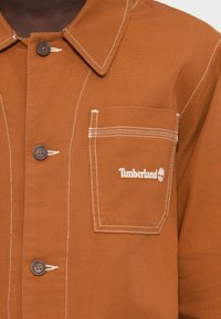 Veste-chemise marron avec fermeture à boutons, surpiqûres blanches contrastantes et poche poitrine gauche brodée du logo Timberland.