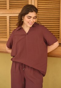Jeune femme portant une chemise ample couleur bordeaux et un pantalon, souriante et regardant vers le bas, debout contre des volets en bois à lattes et un mur jaune.