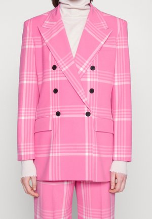 Kobieta ubrana w jasnoróżową, dwurzędową kratkowaną marynarkę z czarnymi guzikami i pasujące spodnie, na białym golfie.