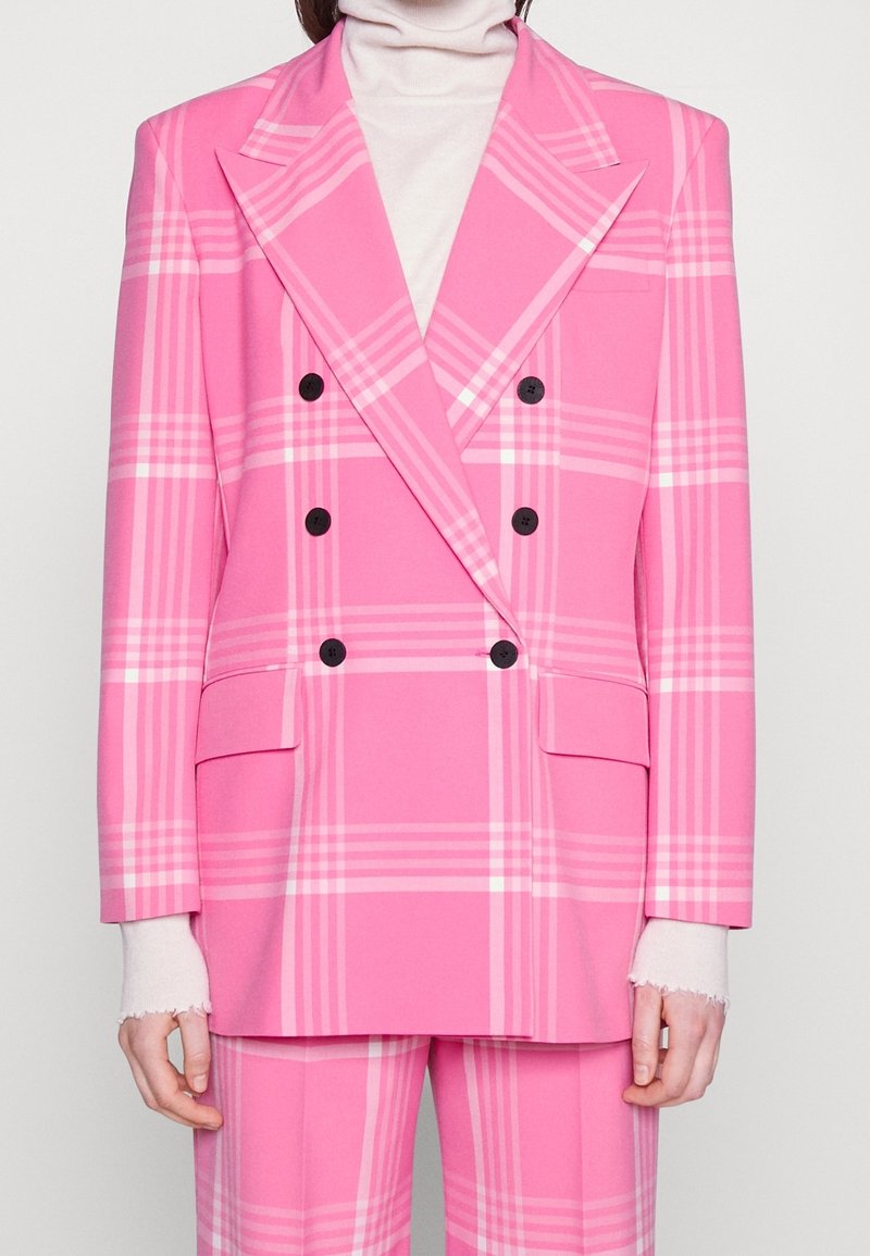 Femme portant un blazer croisé à carreaux rose vif avec des boutons noirs et un pantalon assorti sur un pull à col roulé blanc.