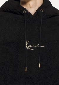 Felpa nera in ecopelliccia con cappuccio e hardware dorato, caratterizzata da una firma decorativa bianca sul davanti e lacci neri.