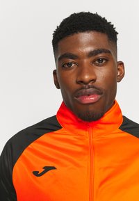 Oranžs un melns sporta žakets ar augstu apkakli, rāvējslēdzēja aizdari un logo uz krūtīm. Gluda auduma virsma ar kontrastējošām krāsām.