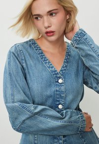 Kék farmerruha V-nyakkivágással, hosszú ujjakkal és ezüstszínű gombokkal. Az anyaga texturált felületű, és enyhén derékban szabott.