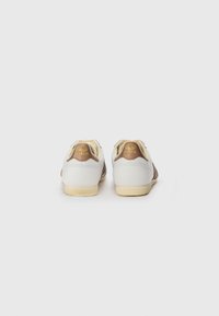 Adidași albi cu accente maro, talpă din cauciuc texturat și logo auriu pe călcâi. Design simplu, cu linii curate și fără embellishments suplimentare.