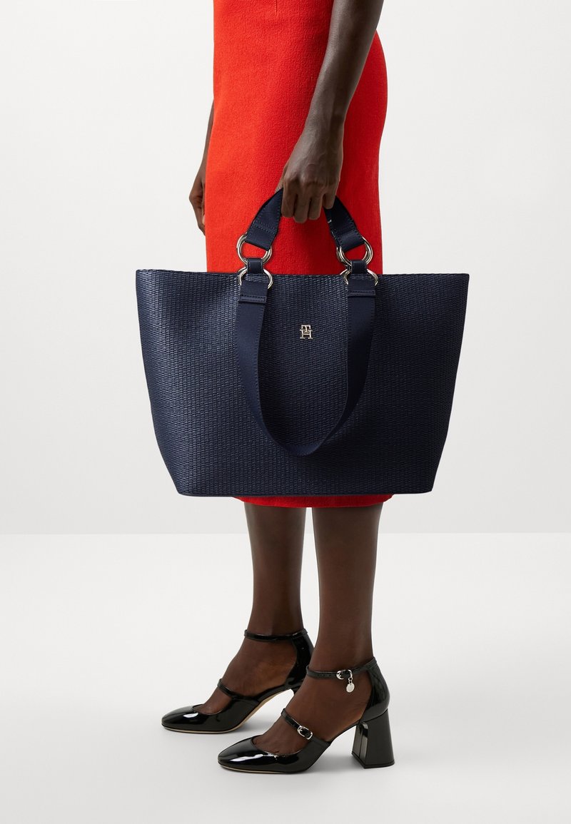 Personne en robe rouge tenant un grand sac à main texturé bleu marine, portant des chaussures à talons carrés en cuir verni noir avec des brides à la cheville.