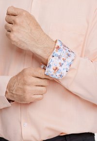 Camisa de tono rosa claro, con textura y un puño con patrón floral. El puño presenta flores azules y naranjas sobre un fondo blanco.