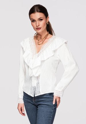 Frau mit dunklen Haaren trägt eine weiße Bluse mit Spitzenbesatz und Volants sowie blaue Jeans, dazu eine goldene Halskette und Creolen-Ohrringe.