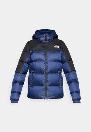 Modra in črna puffer jakna s kapuco, ki ima zadrgo na sprednji strani, dve sprednji žepi in logo na zgornji levi strani. Tekstilirana tkanina.