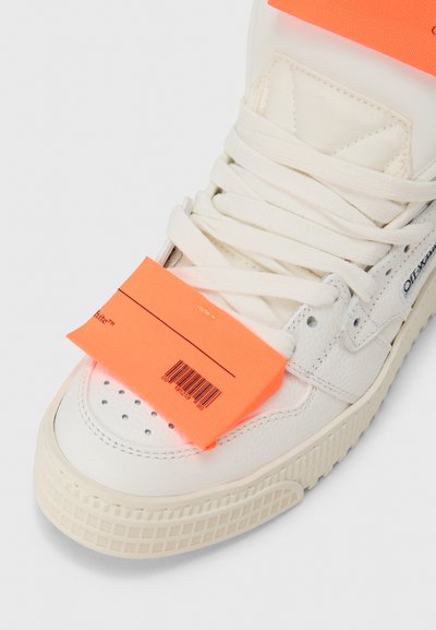 Άσπρο δερμάτινο αθλητικό παπούτσι με υφή στην επιφάνεια, επίπεδα λευκά κορδόνια και έντονη πορτοκαλί ετικέτα με γραμμωτό κώδικα προσαρτημένη στη γλώσσα.