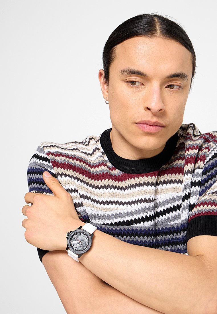 Pull en laine avec un motif en zigzag en crème, gris, bordeaux et bleu marine ; col en côtes noir. Montre blanche avec un cadran circulaire et des détails noirs.