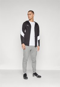 Czarna i biała bluza z kapturem na zamek z logo, w połączeniu z szarymi spodniami dresowymi. Model nosi czarne buty sportowe; prosty, casualowy design.