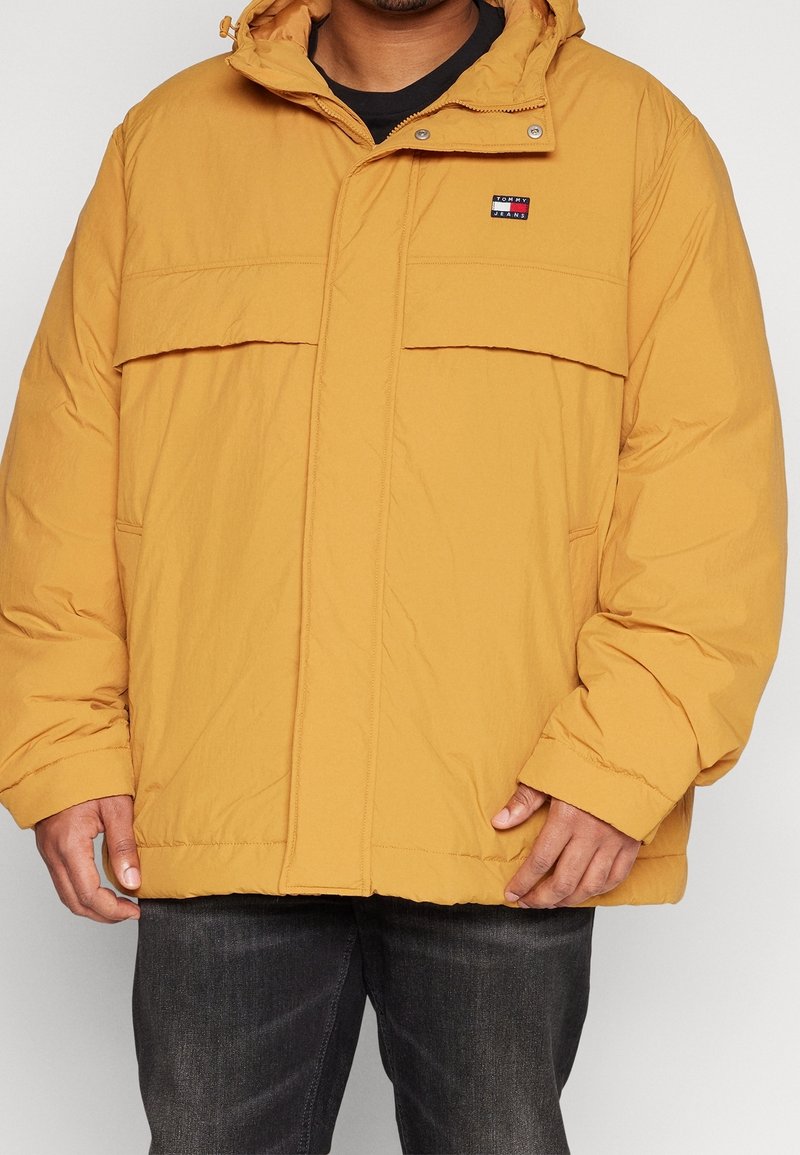 Senapsgul puffjacka med huva, som har en frontdragkedja, två stora fickor med klaff och en liten logopatch på bröstet.