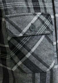 Detalle de camisa de franela a cuadros grises que presenta un bolsillo en el pecho, cierre de botones y una tela suave y texturizada con patrones en blanco y negro que se cruzan.