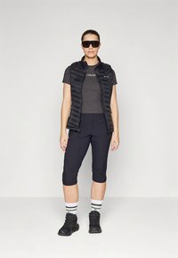 Toppitud must vest tumeda halli t-särgi kohal, sellega koos põlveni ulatuvad mustad püksid, triibulised sokid ja tumedad kingad. Stiili täiustavad päikeseprillid.