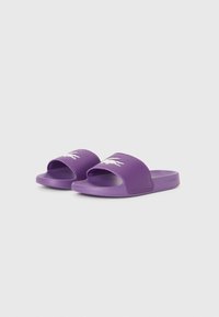 Violetiniai šlepetės su plačiomis juostelėmis, ant kurių pavaizduoti balti krokodilo logotipai, tekstūruotais padu ir šone įspaustu žodžiu "Lacoste".