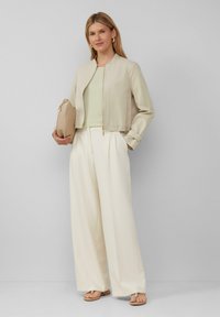 Beige geschnittene Jacke mit Reißverschluss, hellgrünes Oberteil und weite crèmefarbene Hosen. Die Accessoires umfassen eine sandfarbene Clutch und Sandalen mit einem schlichten Design.