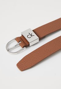 Hnědý kožený opasek Calvin Klein s texturovaným povrchem, stříbrná kovová spona se znakem a vyrytým názvem značky na pásku.