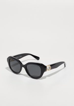 Schwarze Sonnenbrille mit übergroßen, eckigen Rahmen, grauen Linsen und einem goldfarbenen Akzent mit einem Kristalldetail an der Fassung.