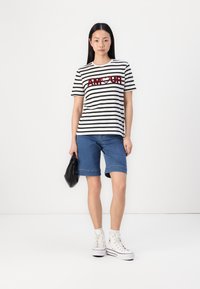 Stripete T-skjorte i svart og hvitt med rød "AMOUR" tekst, parret med denimshorts og høye hvite sneakers. Holder en svart veske.