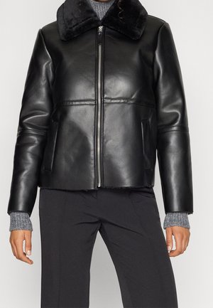 Veste en cuir noire avec fermeture éclair frontale et large col portée sur un pull en maille gris et un pantalon noir.