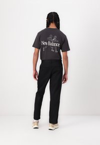 T-shirt gris à manches courtes avec un logo "New Balance" blanc et un graphisme au dos, associé à un pantalon noir et des baskets beige.