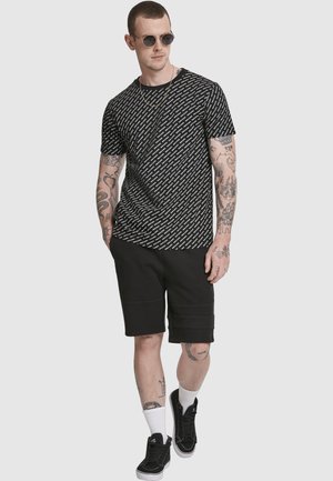 Muž s okrúhlymi slnečnými okuliarmi, v čiernom tričku s vzorom, čiernych kraťasoch, bielych ponožkách, čiernych teniskách, s tetovanými rukami a nohami, stojaci na sivom pozadí.