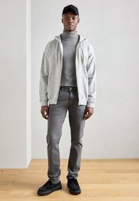 Felpa grigia con zip sopra un dolcevita grigio, abbinata a jeans grigi e scarpe nere. Ambientata su uno sfondo neutro e un pavimento in legno.