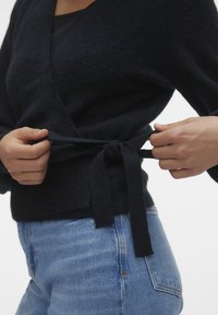 Mörk stickad omslagströja med V-ringning och midjeband, ihopparad med ljusblå denimjeans. Mjuk textur, avslappnad passform.