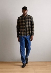 Cămașă cu carouri, în nuanțe de măslin și negru, suprapusă peste un tricou verde; asociată cu blugi albaștri și bocanci din piele neagră.