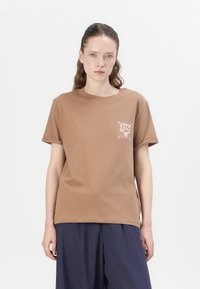 Brown cotton t-shirt with short sleeves, crew neck, and a small graphic print on the left chest. Relaxed fit, smooth texture.