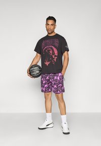 Sort grafisk t-shirt med pink kranie-design, parret med lila mønstrede shorts med kranier. Modellen holder en basketball og har hvide sko på.