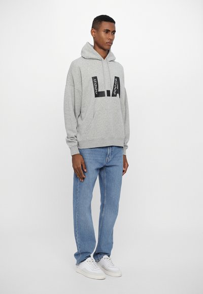 Grauer Kapuzenpullover mit einer Vordertasche und einem schwarzen Logodetail. Kombiniert mit hellblauen Jeans und weißen Sneakern.
