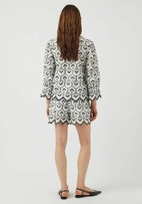Dettagli di ricamo su un romper in tessuto bianco con motivi floreali neri, maniche corte, bordi scallopati e pantaloni coordinati. Scarpe nere flat.