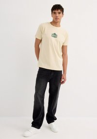Camiseta de algodón beige con diseño de hoja verde en el pecho, combinada con jeans negros y zapatillas blancas. Modelo de pie contra un fondo neutro.