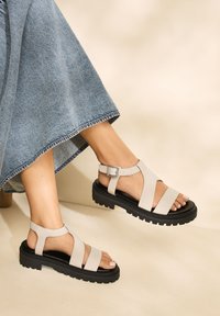 Lyse beige l ædersandaler med justerbar ankelrem, tyk sort gummisål og brede remme, der danner et åbent design. Tekstureret stofkant i baggrunden.