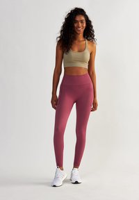 Soutien-gorge de sport beige clair associé à un legging taille haute, extensible, de couleur rose profond. Présence de poches latérales et de chaussures de sport blanches.