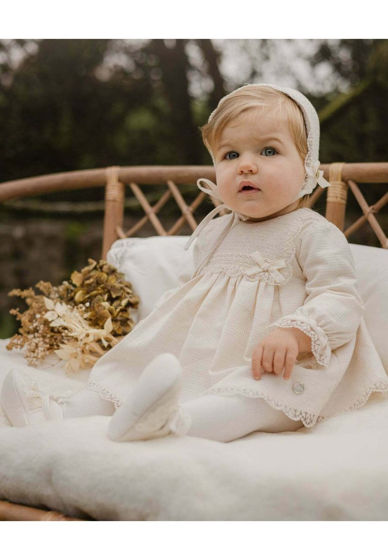 Abito chiaro in crema con tessuto testurizzato e dettagli in pizzo, abbinato a scarpe bianche e collant. Il bambino indossa un cappellino coordinato ed è seduto su una superficie morbida.