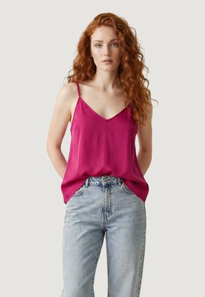 Donna dai capelli rossi che indossa un top magenta senza maniche e jeans azzurri a vita alta, in piedi con le mani dietro la schiena.