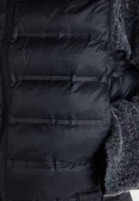Zwarte gewatteerde vest met horizontaal gestikte secties en een grijze wolachtige stof bij de kraag en zijkanten. Voorzien van een rits sluiting en zakken.