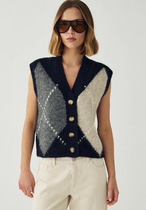 Donna che indossa occhiali da sole marroni oversize e un gilet senza maniche blu navy con bottoni e motivi a rombi grigi e beige.