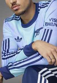 Adidas lys blå sportsjersey med marineblå accenter, stribede ærmer og en klublogo. Blødt stof med en behagelig pasform og ribbet krave.