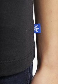 Svart t-shirt med en blå Adidas-tag som har en vit trefoil-logotyp, som visar tröjans struktur och nätt passform längs ärmen.