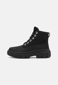 Des bottes de cheville en suede noir avec une semelle en caoutchouc épaisse, un bout rond, des œillets en métal et une languette à l'arrière pour un enfilage facile.