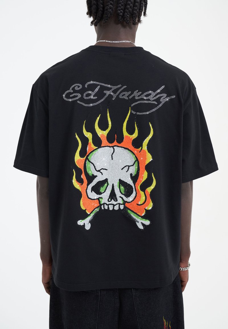 Musta t-paita, jossa on suuri, värikäs pääkallo- ja luurankokuva, koristeena strasseja ja "Ed Hardy" -teksti ylhäällä.
