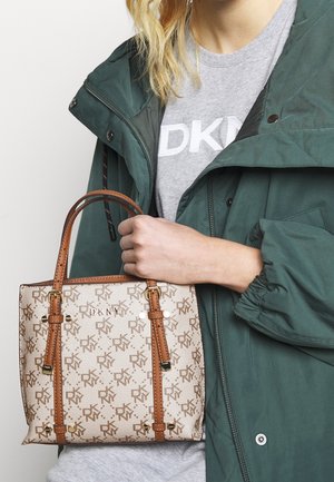 Nainen, jolla on päällä vihreä takki ja harmaa DKNY-paita, pitää käsissään beigeä ja ruskeaa DKNY-monogrammikässä.